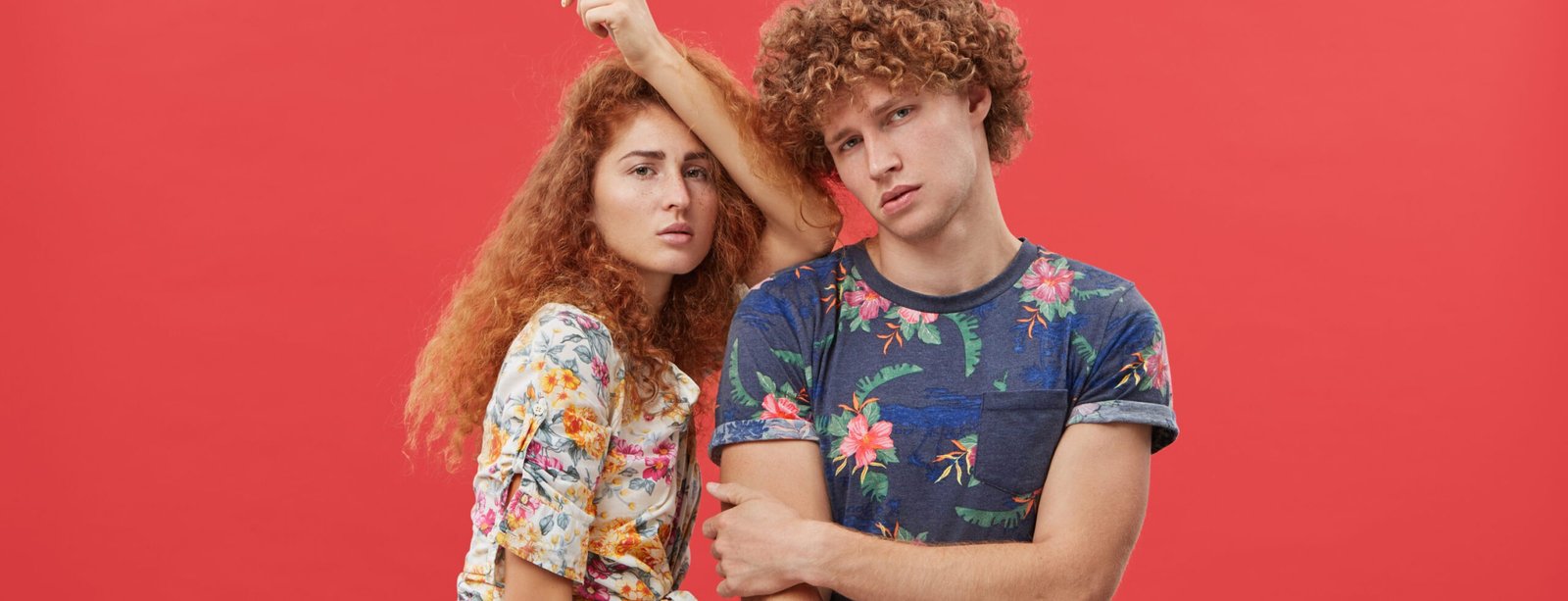 Beautiful woman with red hair and freckles wearing shirt and shorts leaning at shoulder of her curly boyfriend who is posing looking directly into camera with serious expression. Two best friends