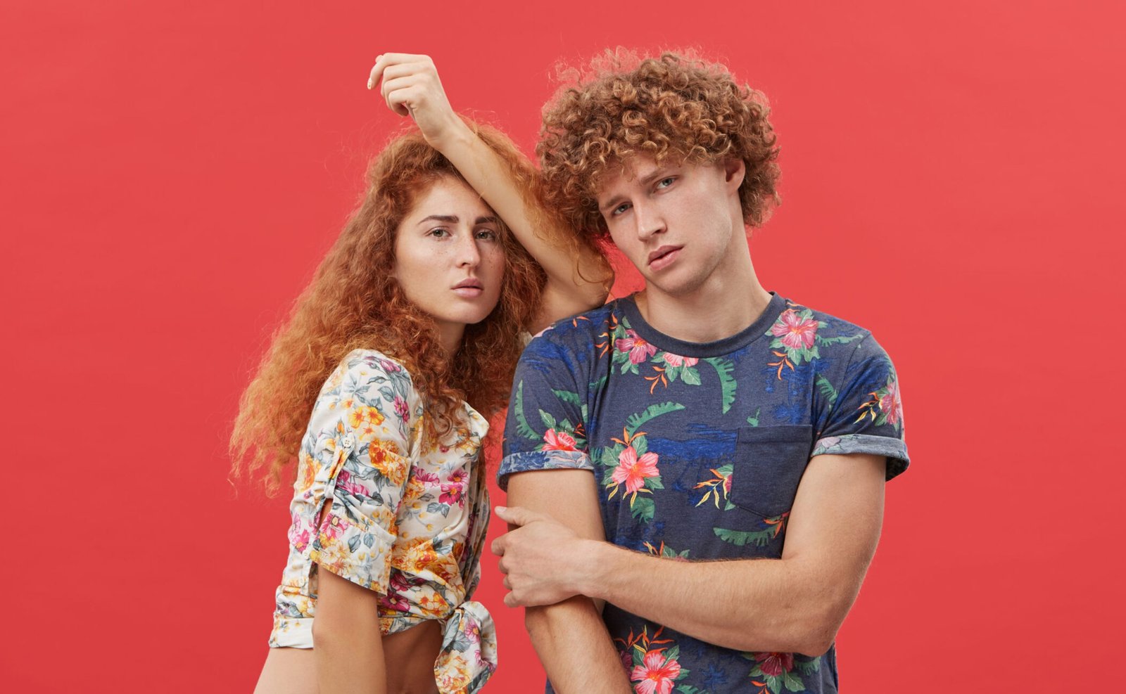 Beautiful woman with red hair and freckles wearing shirt and shorts leaning at shoulder of her curly boyfriend who is posing looking directly into camera with serious expression. Two best friends